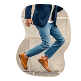 A male model in blue denim pants and a suit jacket jumps in his tan leather shoes by Amberjack.